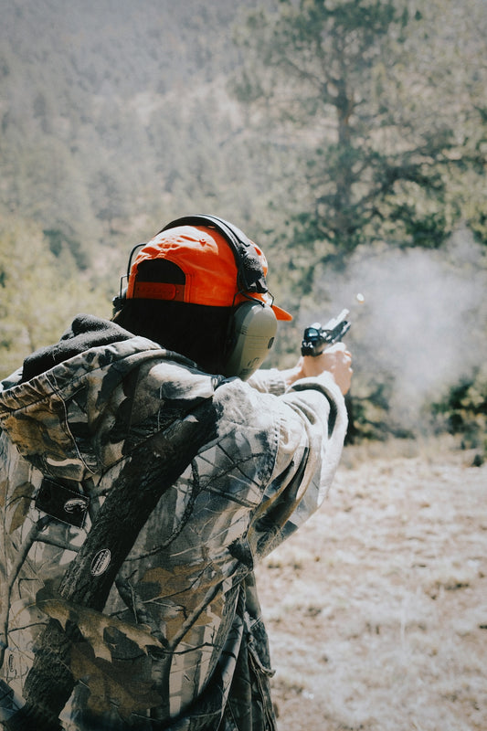 Person in camouflage firing a handgun outdoors with muzzle flash.