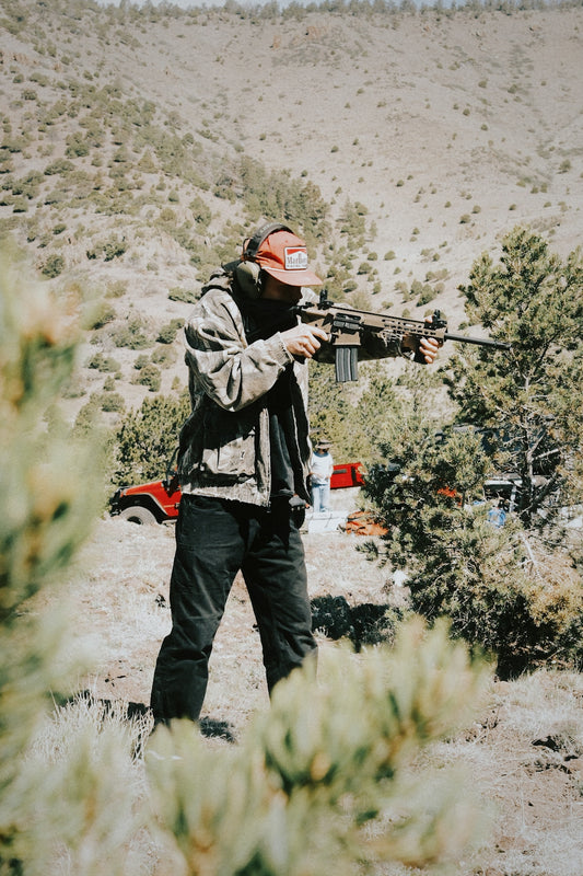 Man shooting a rifle in a dry, hilly landscape.